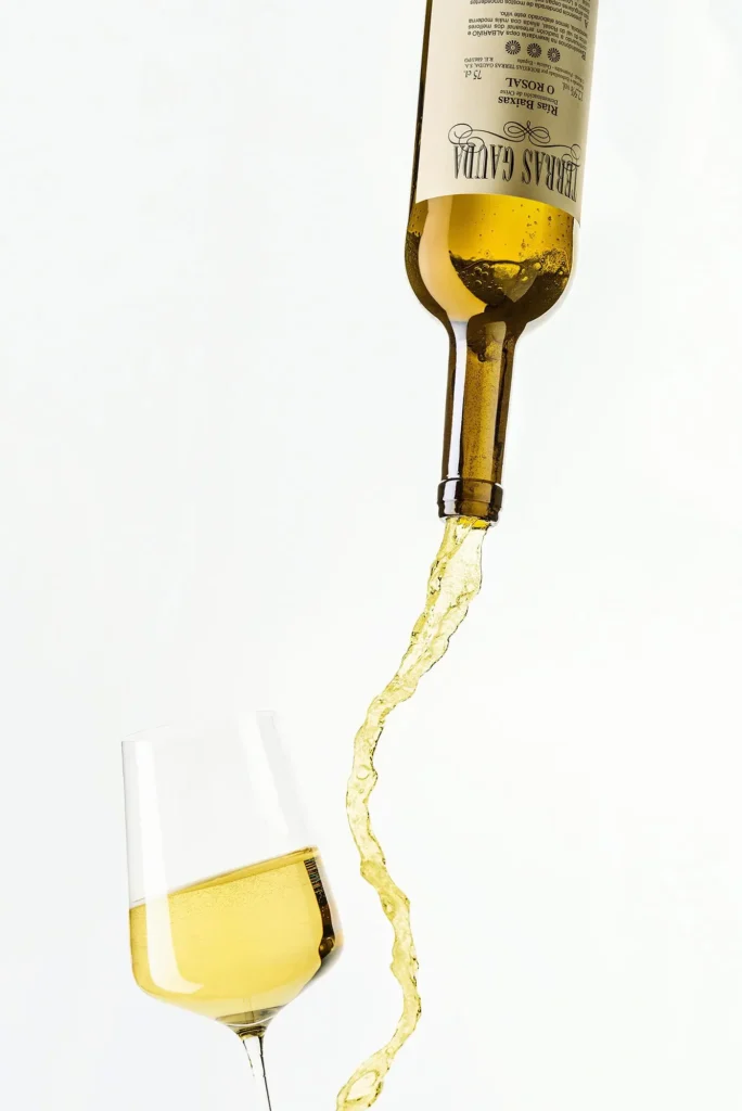 Weissweinflasche mit dem Hals nach unten. Der Wein fliesst schwungvoll aus der Flasche. Am unteren Rand ist ein gefülltes Weinglas.