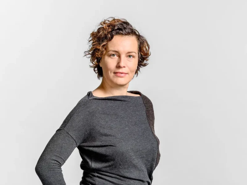 Businessportrait einer Frau mit lockigem Haar im grauen Oberteil vor hellem Hintergrund