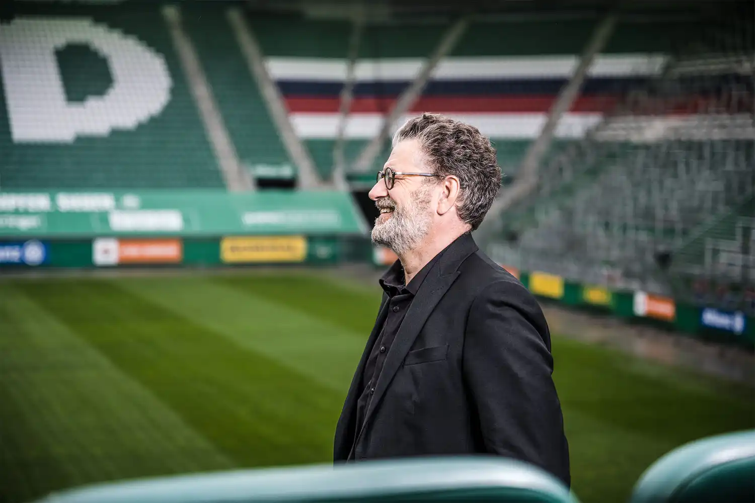 Businessportrait – Architekt Guido Pfaffenhausen in schwarzem Anzug mit Brille lächelnd am Spielfeldrand im Rapidstadion Wien