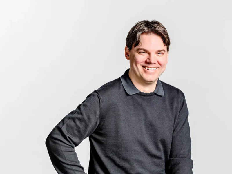 Businessportrait eines Mannes mit dunklem Pullover und breitem Lächeln vor hellem Hintergrund