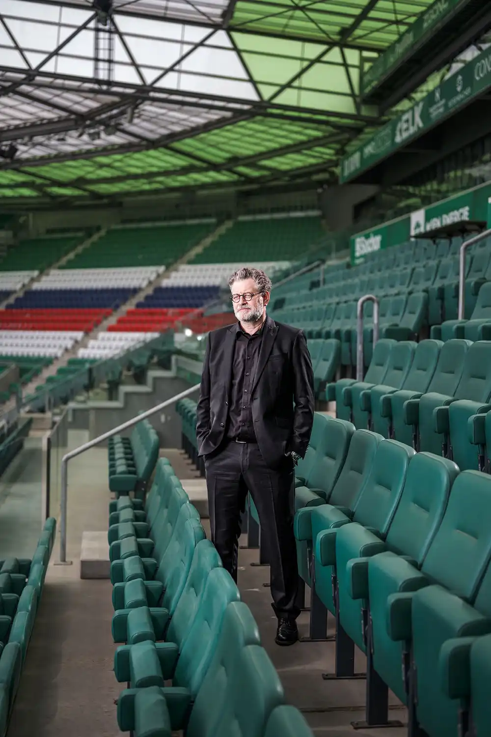 Businessportrait – Architekt Guido Pfaffenhausen in schwarzem Anzug mit Brille zwischen Stadionsitzen im Fußballstadion frontal