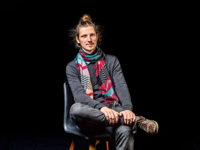Künstlerportrait eines Mannes mit Dutt und buntem Schal auf schwarzem Stuhl vor schwarzem Hintergrund