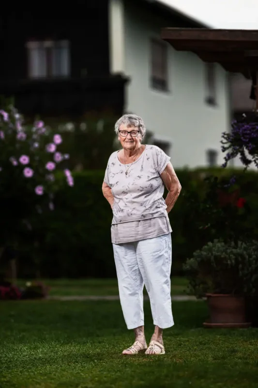 Portraitfotografie – Ältere Frau mit Brille in hellem Sommeroutfit im Garten vor österreichischem Haus
