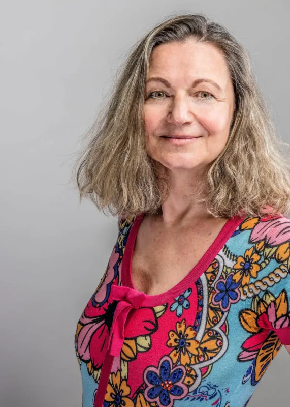 Portraitfotografie – Frau mit grau-blondem Haar in buntem Blumenshirt, leicht seitliche Pose vor hellem Hintergrund