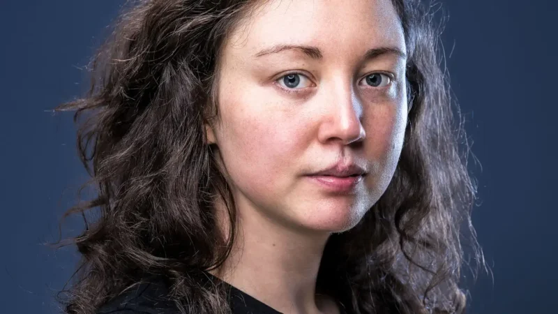 Portraitfotografie – Nahaufnahme einer jungen Frau mit lockigem braunem Haar und blauen Augen vor dunkelblauem Hintergrund