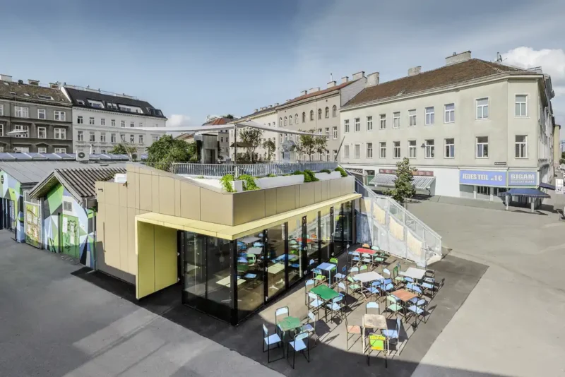 Professionelle Architekturfotografie des Café Wirr am Brunnenmarkt im 16. Bezirk Wien, Planung A2K Architekten, moderner Pavillon mit Glasfassade, Dachterrasse mit Begrünung und Außenterrasse mit bunten Stühlen, fotografiert von Martin Croce Wien